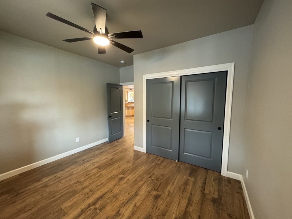Modern remodeled bedroom with wood-look flooring, gray sliding closet doors, neutral walls, and ceiling fan lighting, opening into a hallway toward a kitchen area.