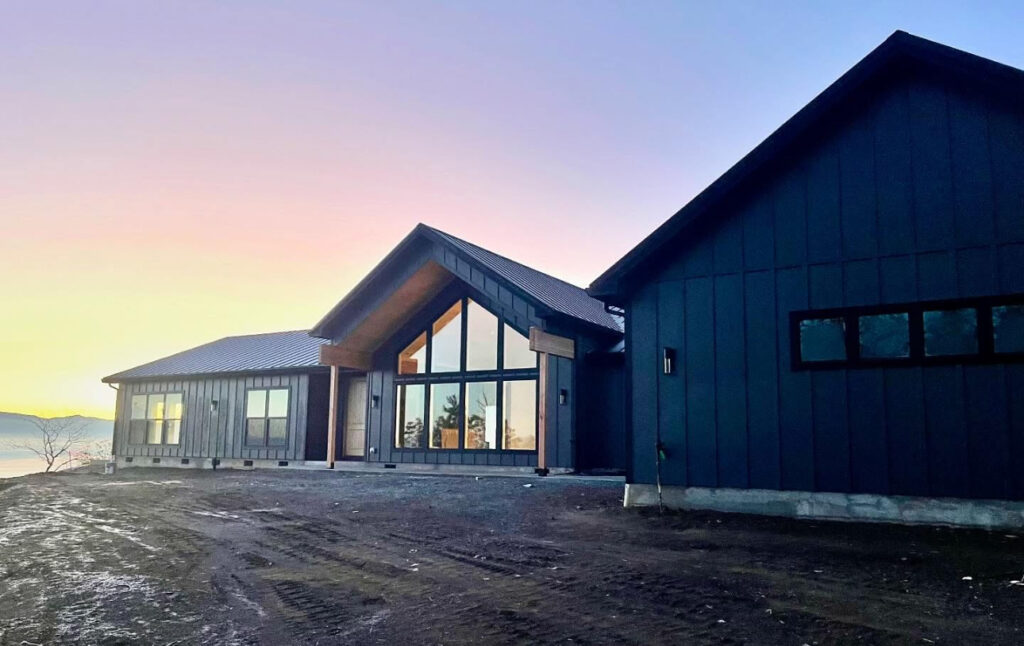 Modern custom home under construction in Southern Oregon with dark siding, large windows, and a dirt-graded lot at sunset